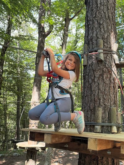 A young girl is depicted mid-air, suspended by a harness and ropes, participating in an adventure park activity. The image is oriented sideways, with the girl looking directly at the camera with a cheerful expression. She is wearing a light blue helmet, a white short-sleeved t-shirt with a graphic design, grey leggings, and grey and pink sneakers. Her body is angled, and her hands are holding onto a metal mechanism attached to a thick metal cable.

The scene is set outdoors, likely in a forest or treed area, suggested by the numerous tree trunks and green foliage visible in the background. A large tree trunk, which appears to be part of the adventure course structure, runs horizontally across the upper part of the image, with wooden planks and metal cables attached to it. Another wooden structure, possibly a platform, is visible to the right of the girl, also integrated with the tree. The sunlight filtering through the leaves suggests it is daytime with clear weather.

The girl's harness, secured around her waist and thighs, and the associated safety equipment, indicate she is engaged in a climbing or ziplining activity. Her smile conveys enjoyment and excitement. One of her sneakers shows the Adidas brand logo with three stripes. The activity appears to be taking place high above the ground, given the perspective looking down through the trees.