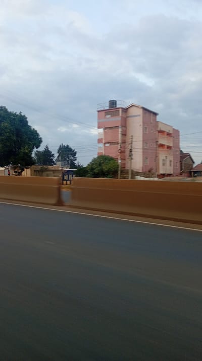 A building with a water tank on top stands behind a concrete barrier. The building has a pinkish hue with lighter accents. Trees are visible to the left of the building. Power lines stretch across the sky. The road is dark and smooth. The sky is filled with clouds, suggesting overcast conditions.