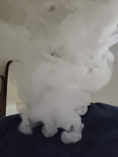 A thick cloud of white vapor dominates the frame, swirling and obscuring what lies behind. To the right, a dark blue fabric contrasts sharply with the vapor. A glimpse of a white surface and a dark wooden frame can be seen through gaps in the cloud, suggesting a background setting. The vapor appears dense and voluminous, creating a soft and diffused effect.