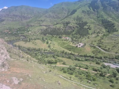 This is an aerial view of a mountainous landscape in Vardzia, Georgia. The scene depicts a lush green valley with a winding river, dotted with trees and scattered buildings. In the distance, a large mountain dominates the background, its slopes covered in vegetation and rocky outcrops. The time of day appears to be daytime, with bright sunlight casting shadows and illuminating the terrain. The weather seems to be clear and pleasant.

The valley floor features a river flowing through a green expanse of fields and shrubbery. Along the riverbanks, numerous trees form a dense, verdant border. Several small clusters of buildings, resembling a village or a small settlement, are visible amidst the greenery, particularly on the left side of the valley. A road winds its way through the landscape, connecting different areas and leading to a parking area where a bus is parked. The terrain in the foreground is characterized by a grassy slope interspersed with rocks and boulders, suggesting a natural, undeveloped environment.

The overall impression is one of a serene and picturesque rural setting, characterized by the natural beauty of the mountains and the river valley. The presence of a road and buildings indicates human habitation and infrastructure within this natural environment. No people are clearly visible in the image. There is no discernible text in the image.