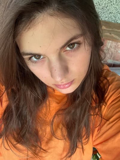 A close-up, high-angle selfie shows the face of a young woman with long, dark brown hair. She is looking directly at the camera with a neutral expression. Her eyes are brown, and she has light skin with a few freckles. She is wearing an orange long-sleeved shirt. The background is blurred, but appears to be an outdoor setting with some wooden planks and a textured surface. The scene appears to be set in Tsarski Izvor, Bulgaria.