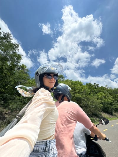 Two individuals are riding on a motorcycle on a paved road surrounded by lush green trees under a partly cloudy blue sky. The person in the foreground, wearing a light-colored, sheer blouse, denim shorts, and a grey helmet with a clear visor, is looking to the side. A colorful scarf is tied around their neck and flutters in the wind. They are wearing sunglasses and appear to be taking a selfie. The second person, in the background, is wearing a pink t-shirt and a black helmet and is operating the motorcycle. The road has yellow markings. The scene suggests a journey through a rural or natural area, possibly in the vicinity of Ham Thuan Bac, Vietnam, given the tropical foliage.
