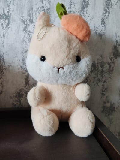 A plush toy rabbit with light peach-colored fur and a white muzzle sits on a dark, flat surface. The rabbit has black button eyes, a small brown embroidered mouth, and a green leafy carrot-shaped hat on its head. A thin silver string loop is attached to the top of the rabbit's head. The background is a textured wall with a mottled gray and white pattern. The lighting is soft, suggesting an indoor setting, likely during the day. There is no visible text in the image. The overall scene is simple and focuses on the plush toy.