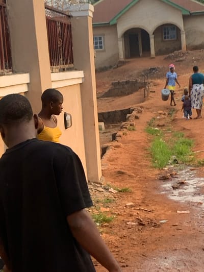 This outdoor scene, set in Otuocha, Nigeria, captures a glimpse of daily life. The ground is primarily reddish-brown earth, uneven in places, with sparse patches of green grass and some lighter, possibly sandy or disturbed, areas.
In the background, a two-story building with light-colored walls and a green roof is visible. It features distinct arched entrances or windows on the ground floor and rectangular windows with white frames on both levels. To the left in the mid-ground, another building with concrete walls and a metal gate/fence is partially seen. A dirt path or slope with rough steps or retaining structures appears to lead down.
Several people are present. In the far background, a woman wearing a patterned skirt and a dark top is seen holding a small child by the hand. Further back on the left, another person, likely a woman, is walking while balancing a light grey or white bucket on her head. She is dressed in a light blue top, a yellow skirt, and a pink head covering. In the foreground on the right, a person with short dark hair, wearing a black t-shirt, is seen from the back and side. Slightly behind them, another individual with short hair, wearing a yellow top, is shown in profile, appearing to look upwards or to the side.
The scene depicts common activities like walking and carrying goods, suggesting routine daily life in a residential or semi-rural setting. The natural lighting indicates it is daytime, possibly late afternoon, with no harsh shadows. No text is visible in the image.