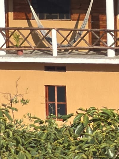 An exterior shot reveals an orange building partially obscured by dense green foliage on the right. A window with a dark interior and wooden frame is visible on the building. To the left, an architectural detail of what appears to be a balcony is framed by a white support structure with a diagonal metal latticework pattern. A hammock or awning is partially seen, suggesting a relaxed outdoor space.