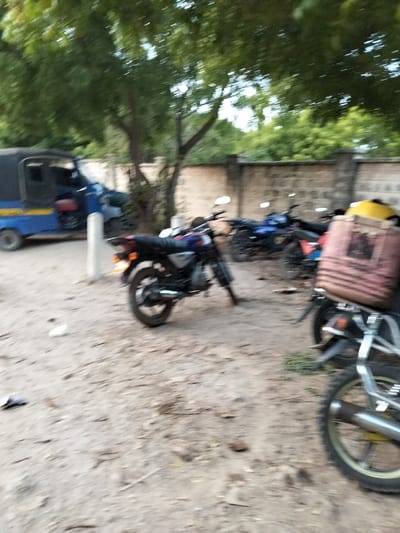 This image captures an outdoor scene in Gombato Bongwe ward, Kenya, featuring an unpaved dirt parking area under a canopy of trees. A light-colored block wall forms the background. In the mid-left, a blue and yellow three-wheeled auto-rickshaw is parked. Multiple motorcycles are stationary on the dirt ground. A dark-colored motorcycle with chrome elements is visible in the mid-left foreground, parked on its kickstand. To its right, another motorcycle carries a light brown and pink striped bag on its rear rack, with a yellow object behind it. Additional motorcycles, including a blue one, are visible further back towards the wall. A section of a motorcycle's front wheel and exhaust pipe are present in the immediate right foreground. No individuals are discernible in the scene.