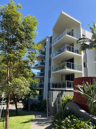 The image displays a modern, multi-story apartment building under a clear, vibrant blue sky. The building's facade is predominantly light-colored, possibly off-white or light gray, complemented by darker gray and reddish-brown architectural accents. It features a distinctive design with multiple balconies characterized by prominent, sharp-angled overhangs and extensive glass railings and windows. These glass surfaces reflect the bright sky and the lush green trees surrounding the structure.
In the foreground and around the base of the building are numerous trees with dense green foliage, suggesting a well-landscaped environment. A large palm frond is visible near the top of the building, and other tropical plants are integrated into the landscaping along the building's perimeter. The ground level includes patches of well-maintained green grass and a paved pathway. A temporary metal fence stands near the pathway and the lower section of the building, hinting at ongoing work or restricted access.
The scene is captured during bright daylight, with clear skies and distinct shadows indicating mid-morning or afternoon. There is no visible human activity or discernible text in the image. The setting appears to be a contemporary residential or mixed-use area in Kelvin Grove, Australia, emphasizing the architecture and natural elements.