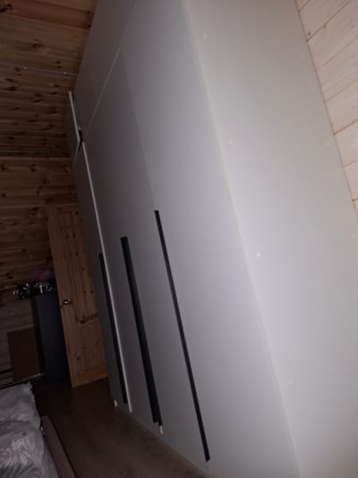 The image shows a large, modern, light gray wardrobe with dark handles, positioned in a room with wooden walls and ceiling. The wardrobe doors are sleek and minimalist, with vertical dark lines serving as handles. One door on the left appears slightly ajar, revealing a sliver of its interior. To the left of the wardrobe, a wooden door with a round knob is visible, partially obscured by a dark gray cabinet or appliance. On top of this cabinet, there are several metallic objects, possibly containers or canisters. The floor is made of dark wood. The overall impression is of a functional and contemporary interior, likely a bedroom or living space. The location is identified as Medvezhyi Ozyora, Russia.