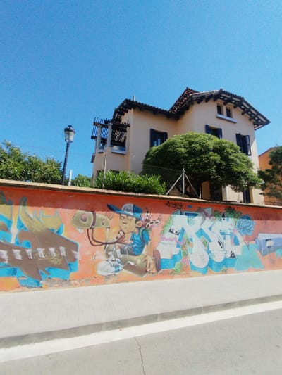 The image captures a vibrant street scene in Rubí, Spain, under a clear, bright blue sky, suggesting a sunny day. In the foreground, a light-colored paved sidewalk runs parallel to a darker asphalt road, separated by a curb and a white painted line.
Dominating the mid-ground is a long, orange-painted wall extensively covered in graffiti. The graffiti features a variety of colorful designs, including abstract shapes, stylized letters, and several character depictions. A prominent character on the left side of the wall appears to be a young man or boy wearing a blue baseball cap, holding a leash attached to a light green, four-legged creature with a large, elongated snout. Other graffiti elements include a large blue letter-like design and what resembles a blue flower on the right. Some visible text within the graffiti includes "RAZ" or "PAZ" on the left.
Behind the graffiti wall, a substantial multi-story building with a light beige or cream facade and a red-tiled roof is visible. The building features dark blue window shutters and decorative wooden eaves or beams under the roofline. Green trees and bushes are planted around the building, with a particularly large, round-canopied tree standing centrally in front of it. A chain-link fence is also discernible between the wall and the building, partially hidden by the foliage. To the left, a traditional black street lamp with an ornate top stands tall, positioned between the graffiti wall and the building's roof.