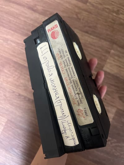 The image shows a close-up of two VHS tapes lying on a wooden surface. The tapes are black and have labels with handwritten text. The tape in the foreground has a white label with text written in Cyrillic, appearing to be a title and possibly names. The tape in the background has a lighter label with some red graphic and printed text in Cyrillic, indicating it is from RAKS and shows production information including the year 1995. A hand is partially visible, holding the tapes. The background is a wooden floor with a distinct grain pattern. There is no visible indication of the city of Amirovo, Russia, or any other specific location.