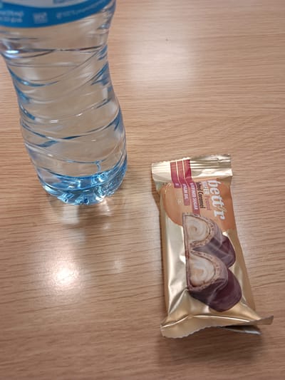 The image shows a close-up, overhead view of a plastic bottle of water and a golden foil-wrapped snack on a wooden surface. The water bottle is clear with blue accents and appears to be partially filled. The snack is labeled "bettr" and displays text that includes "Wafer Peanut," "Salted Caramel," and "UNREFINED SUGAR." A portion of the snack is visible through the packaging, revealing layers of wafer and caramel. The wooden surface has a visible grain pattern. There are no people visible in the image. The scene is static and appears to be a simple arrangement of food and drink on a table. The location is not specified within the image itself, and there is no contextual information provided that would link it to Radinovo, Bulgaria.