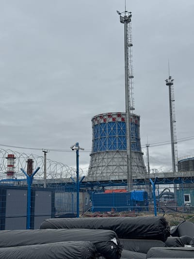 In the city of Chulman, Russia, a industrial scene unfolds under an overcast sky. The foreground is dominated by large, black, rolled-up materials, possibly for construction or insulation, stacked and secured. Behind them, a blue chain-link fence topped with barbed wire signifies a secure perimeter. Mounted on blue posts are security cameras, actively monitoring the area.

Beyond the fence, two industrial cooling towers stand prominently. The larger one, in the center, is a massive cylindrical structure with a distinctive blue lattice pattern on its upper section, accented with red and white. It is supported by a tall metal structure with a ladder extending to its peak, which is topped with lights. To its right, another, smaller cooling tower or industrial chimney with a red and white striped top is visible. A second, taller metal tower with lights and a ladder also stands to the right of the main cooling tower, its peak silhouetted against the sky. In the distance, additional industrial structures and tanks are partially obscured by the haze and the barbed wire fence. The overall environment appears to be a working industrial site, possibly undergoing construction or maintenance, given the presence of the rolled materials.