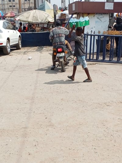 This image captures a daytime street scene in Naze, Imo State, Nigeria, characterized by an unpaved, light brown dirt road with visible tire impressions and scattered debris.
In the midground, an adult male, appearing to be of African descent, is seated on a dark motorcycle, facing away from the viewer. He wears a multi-patterned long-sleeved sweater (incorporating grey, black, white, and brown) and dark trousers. The motorcycle displays a red taillight and a Nigerian license plate, "IMO KQD-127QZ." To the immediate right of the motorcycle, a young boy, also of African descent, walks rightward, balancing a green plastic crate filled with clear plastic sachets (likely containing water) on his head. He is dressed in a dark grey t-shirt, ripped light blue denim shorts, and dark flip-flops.
The background reveals a bustling commercial environment. On the far left, a white SUV is partially visible, parked. Behind the motorcycle, a market area features several stalls, identifiable by large white and smaller colorful umbrellas providing shade. A person in an orange garment is partially visible under a prominent white umbrella. Further to the right, a blue metal barrier with gold decorative accents separates the foreground from a brown structure bearing the white text "POS" and "AVAILABLE," indicative of a Point of Sale service. A figure in dark clothing is discernible behind this barrier. Shadows cast on the dusty ground suggest bright, direct sunlight from the left.