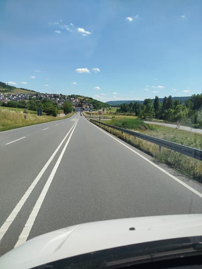 The image shows a view from the driver's perspective of a car on a two-lane road. The road has a white solid line and a dashed white line separating the lanes. In the distance, there are several cars on the road. To the left of the road, there is a hillside with a town nestled on it. The town has many houses with red roofs and is surrounded by trees. To the right of the road, there is a metal guardrail and more trees. The sky is blue with a few white clouds. The location is identified as Distelhausen, Germany.