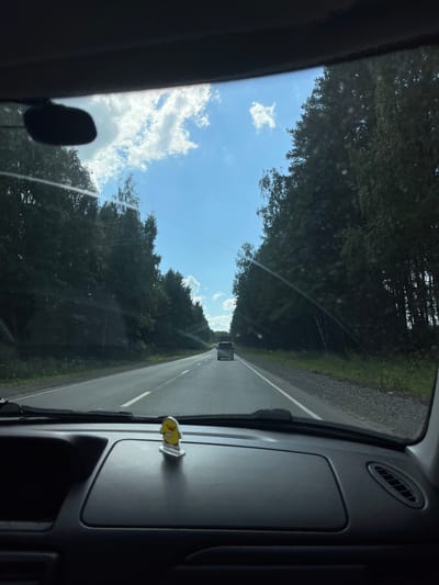 The image is a point-of-view shot from inside a vehicle, looking out through the windshield. The vehicle is on a paved road, traveling through a wooded area. The road is flanked by dense trees on both sides, with a bright blue sky and scattered white clouds visible above. There are two other vehicles visible on the road ahead: a white SUV further down the road and a dark-colored minivan closer to the foreground. On the dashboard of the vehicle from which the photo is taken, a small yellow duck figurine is visible. The scene suggests a rural or semi-rural setting, consistent with a location like Uspenskii, Russia, characterized by roads winding through natural landscapes.