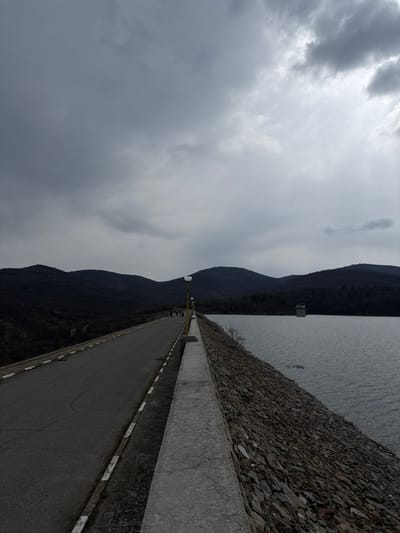 This image depicts a wide-angle view along the crest of a dam, located near Баня, Bulgaria, under an overcast sky. A two-lane asphalt road occupies the left side of the frame, extending into the distance and bordered on its right by a solid white line and a broken yellow line. Parallel to the road, a low, light-grey concrete barrier separates it from the dam's edge. Mounted atop or behind this barrier is a series of evenly spaced yellow lamp posts, each with a white, circular light fixture, receding into the background.
To the right of the concrete barrier, the dam's slope is covered with dark, angular riprap stones that descend into a large body of dark, calm water, which fills the right half of the image. In the mid-distance on the water, a dark, rectangular concrete tower structure is visible. The background features dark, undulating mountains, densely covered with vegetation. The sky is completely overcast with multiple layers of grey clouds. In the far distance on the road, approximately 3-5 indistinct figures, appearing as dark silhouettes, are observed walking away from the viewer. No specific actions or interactions between these individuals are discernible. The scene is dimly lit by diffused daylight.
