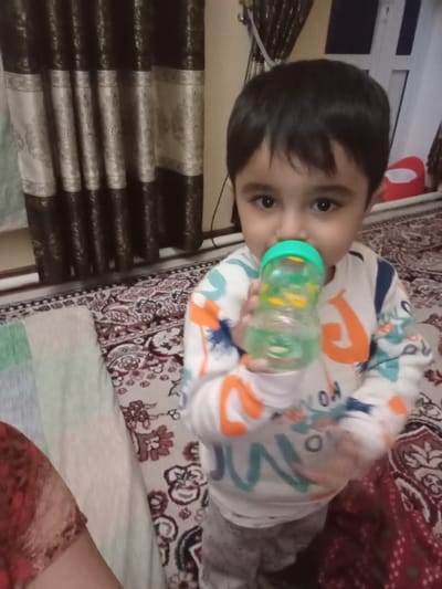 A young child, approximately two to four years old, with dark hair and dark eyes, is depicted from the chest up, positioned centrally in an indoor residential setting in Galaosiyo, Uzbekistan. The child wears a white long-sleeved shirt patterned with multi-colored abstract shapes in orange, teal, and blue, over light gray patterned pants. The child holds a clear plastic sippy cup with a bright green lid, appearing to drink from it; the cup contains a light green liquid. To the lower left, a portion of an adult's arm, clad in a reddish-brown patterned garment, is visible. The background features dark brown curtains with light beige vertical patterned bands on the left, and a partially visible white window or door frame with a red object on the right. The floor is covered by a patterned rug or carpet displaying beige, brown, and red floral motifs.