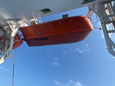 This media file shows a view looking up from below at an orange lifeboat suspended from a ship's davit system. The lifeboat is the primary subject, with its curved hull and orange exterior clearly visible. The davit, a metallic structure designed to lower the lifeboat, is also prominent, supporting the lifeboat with cables and hooks.

The scene is set against a bright blue sky with scattered white clouds, suggesting fair weather during daylight hours. The underside of a white ship deck is visible in the upper portion of the image, with structural elements and what appear to be recessed hatches or openings. Another lifeboat, partially obscured, can be seen to the left of the main subject. The overall impression is one of a maritime setting, likely on a commercial vessel or ferry. There is no visible text in the image. The activity taking place is implied – the lifeboat is in a stowed position, ready for potential deployment. No people are visible.