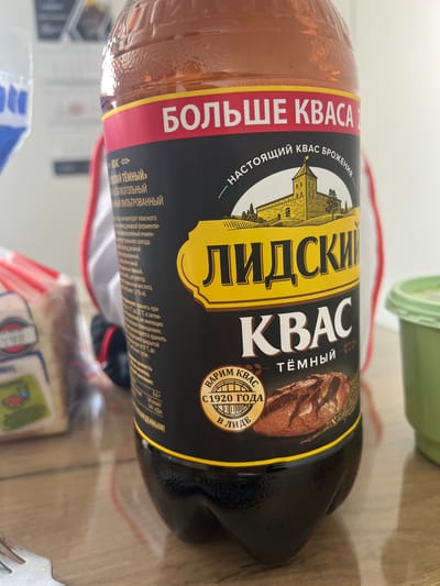 The image shows a close-up of a dark-colored bottle of kvass, a fermented beverage. The label on the bottle is predominantly black with yellow and red accents. It features the brand name "LIDSKI KVASS" in large yellow letters, with "TËMNY" (dark) below it. Above the brand name, there is an illustration of a castle and the text "НАСТОЯЩИЙ КВАС БРОЖЕНИЯ" (real fermented kvass). The top of the label has the text "БОЛЬШЕ КВАСА" (more kvass). There is also a circular emblem with the text "ВАРИМ КВАС С 1920 ГОДА В ЛИДЕ" (we brew kvass since 1920 in Lida), which is a city in Grodno Region, Belarus. The bottle is placed on a wooden table. In the background, out of focus, are glimpses of other items, including what appears to be a loaf of bread in packaging on the left and a green container on the right. The lighting suggests an indoor setting, possibly a kitchen or dining area. The image composition is focused on the kvass bottle. The specific location in Тэльмінскі сельскі Савет is not explicitly indicated in the image.