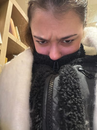 A woman looks into the camera with slightly furrowed brows. She wears a dark jacket with a zipper and fluffy black trim around the collar. There's white fur or stuffing around her shoulders. The photo is taken at an unusual angle, rotated so the woman appears sideways. Background features a wooden shelf unit with books or other items visible.