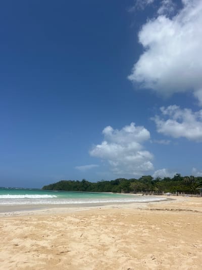 The image displays a wide, unpopulated sandy beach at Drax Hall, Jamaica, under a clear blue sky dotted with white cumulus clouds, primarily clustered on the right. The foreground consists of light beige sand, transitioning to wet sand at the water's edge. Gentle, white-capped turquoise waves break on the shore, extending across the frame from left to right. The ocean water, a vibrant blue-green, stretches towards a distant, heavily vegetated landmass on the left. The coastline in the mid-ground is characterized by a dense treeline of lush green tropical foliage. On the far right, near the treeline, subtle indications of man-made structures, including a white canopy and what appear to be lounge chairs, are partially visible. No individuals, animals, or discernible activities are present. The scene is illuminated by direct sunlight.