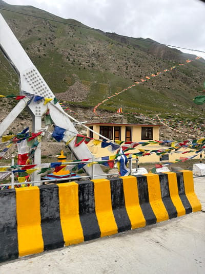 This is a photograph of a scenic mountain landscape in Chikka, India, with a bridge and a building prominently featured. The bridge, constructed with white metal beams and numerous rivets, is adorned with colorful prayer flags. These flags, typically associated with Buddhism, are also strung across the mountainside, creating a festive and spiritual atmosphere. A yellow building with dark-rimmed windows stands near the bridge, also decorated with prayer flags. In the foreground, a concrete barrier painted with alternating black and yellow stripes adds a touch of caution and definition to the scene. The mountains in the background are covered in green grass and rocky terrain, with a cloudy sky overhead. There are no people visible in the image.