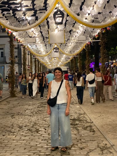 The photograph captures a street scene in Tavira, Portugal, adorned with festive decorations overhead. A canopy of white, petal-like elements and yellow garlands stretches across the cobblestone street, illuminated by strings of lights and decorative lanterns. In the foreground, a woman with dark hair, wearing a white tank top and loose-fitting blue jeans, stands facing the camera, a black crossbody bag slung over her shoulder. She is smiling and her pose is relaxed.

In the background, a diverse group of people are walking along the street. Some are blurred due to motion or distance, while others are more discernible. Their attire varies, suggesting a casual evening outing. The architecture of the buildings lining the street is partially visible, with facades in muted tones. The cobblestone pavement is uneven and scattered with dry leaves or foliage. The overall impression is that of a lively street fair or cultural event at dusk or night.