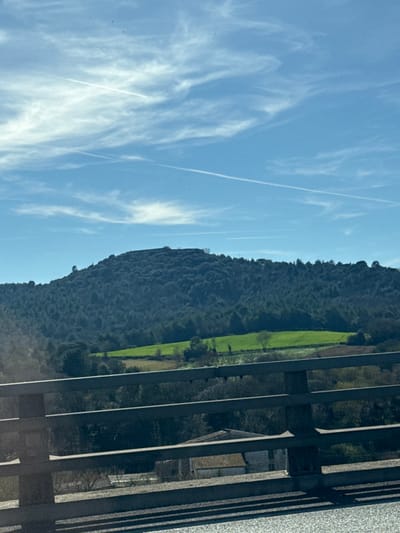 A scenic view presents a mountainous terrain with a vibrant green field alongside it. Overhead, a bright blue sky is decorated with wispy clouds and contrails. The foreground contains a metal railing and building partially obstructing the landscape. The photograph is rotated and taken during daylight.