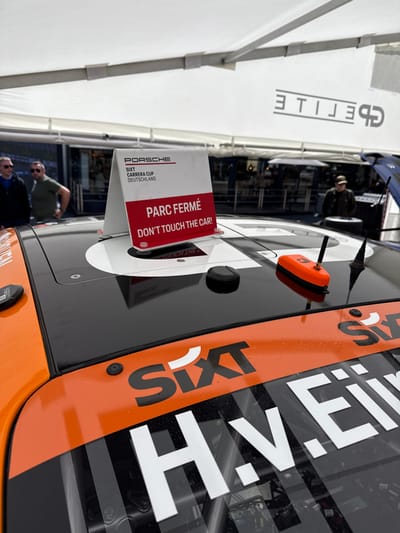 The image shows a black and orange race car with the word "SIXT" printed on the side, parked under a white canopy tent. There is a sign on top of the car that reads "PARC FERME" in red letters, with the phrase "DON'T TOUCH THE CAR!" written below in black. The sign is also labeled with "PORSCHE SIXT CARRERA CUP DEUTSCHLAND." The location is in Stavelot, Belgium, near the Circuit de Spa-Francorchamps, a famous race track. The car appears to be in a closed area for the race. The canopy tent has a large black sign in the background, but it is partially obscured by the tent's fabric and only the word "GP ETT" is visible. There are people in the background, but their details are not clear. The ground is paved. There is a small orange object on top of the car that appears to be a camera. There is also a black object on the car's roof, which is also a camera. The car is parked on a closed racing circuit and the signs indicate it is a race car that has been used. The background is a bit blurry and there are no other details that can be discerned about the environment.