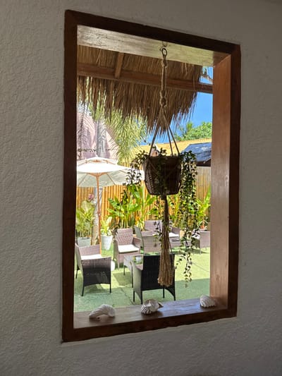 The image displays an outdoor seating area viewed through a rectangular, dark brown wooden window frame from an interior with a textured white wall. Suspended from the top center of the window frame is a macrame-style rope plant hanger holding a woven basket containing a cascading green plant. On the dark brown wooden windowsill, three seashells are arranged: two on the left and one on the right.

The outdoor area features a green ground cover, possibly artificial turf, under bright daylight conditions. Multiple sets of dark brown wicker-style chairs with light cushions are arranged around small tables with glass tops. On the left side, a large white market umbrella is open, providing shade. Behind the seating area, a vertical bamboo fence acts as a partition, with various tropical plants, including broad-leafed species and potted plants, visible. The overhead structure of the outdoor area includes a thatched (nipa) roof. In the background, beyond the bamboo fence and foliage, blue sky is visible, along with distant palm trees and other tropical vegetation, and a darker, partially visible roof structure. The scene is consistent with a casual outdoor lounge or dining area within a commercial establishment in Panglao, Philippines. There are no people present in the visible area.