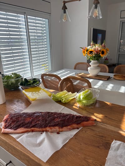 A large piece of salmon is laid on a white piece of parchment paper on a wooden kitchen counter.  It is covered in a thick red sauce.  In the background, there is a white kitchen table with a bouquet of flowers in a white vase.  The table is set with four woven chairs, a white tablecloth, and sunlight streaming across the surface.  On the table, there is a white plastic container of romaine lettuce, an empty container that appears to be yellow, a white paper towel, and a second container of romaine lettuce.  There is another wooden counter that is behind the table with a white paper towel, a white bowl of salad greens, and a container of chopped broccoli.  This scene is located in the city of Gan Yavne, Israel, at the coordinates (latitude: 31.779, longitude: 34.703).