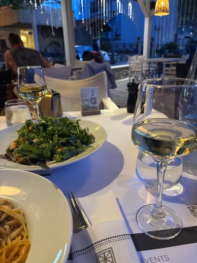 The image depicts a dining scene at an outdoor restaurant in Olympiada, Greece. In the foreground, a white table is set with a plate of pasta, a salad, and two glasses of white wine. A napkin with a patterned edge rests beside the plate of pasta. In the background, a person is visible in the middle distance, facing away from the camera. The restaurant's ambiance is evident, with hanging lights and other tables and chairs visible in the blurred background. The overall setting suggests an evening meal at a coastal eatery.