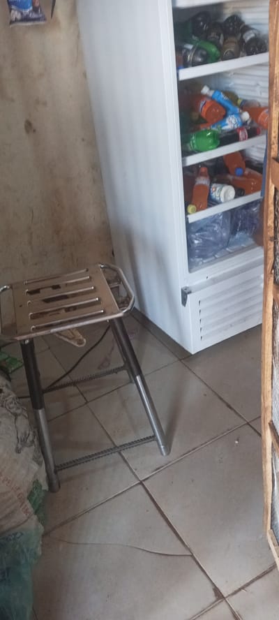 The image captures an indoor scene, likely a storage or small retail area in Ban, Nigeria. A prominent white commercial refrigerator, equipped with a clear glass door, occupies the right side of the frame. Inside, several shelves are filled with various bottled beverages, including sodas, juices, and water, in different colors and sizes. Some bottles are stacked, while others lie on their sides.

To the left of the refrigerator stands a small, metallic stool with a slatted seat and tubular legs. A dark cable or cord is visible near the base of the stool, running across the floor. The floor is made of light-colored square tiles, displaying prominent grout lines, some dirt, and minor cracks.

The wall behind the stool and next to the refrigerator appears stained and dirty, suggesting age or lack of maintenance. A blurry, small image or poster is visible on the upper left portion of this wall. On the far left, part of a large, light-colored sack or bag is visible on the floor. A wooden frame or door jamb is partially visible along the right edge of the image. The scene is static, with no human figures or specific activities depicted. No discernible text is clearly readable on any of the items or surfaces.