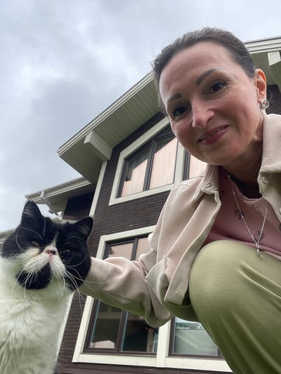 A medium shot captures a woman and a black and white cat in front of a two-story house with dark brown siding and white trim. The woman, appearing to be in her 40s, is crouched down and smiling at the camera, her face illuminated by natural light. She is wearing a light pink t-shirt with a subtle pattern, a beige jacket, and olive green pants. A silver necklace with a pendant rests on her shirt. The cat, a domestic shorthair with distinct black and white markings, is positioned to the left of the woman, looking directly at the viewer. Its fur appears fluffy, and its whiskers are prominent. The background consists of the house's facade, featuring multiple windows with a reddish tint, and a cloudy sky. The perspective is low-angle, emphasizing the subjects from a ground-level viewpoint.