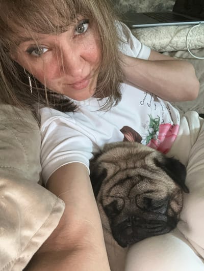 A woman with blonde hair and blue eyes is lying down with a pug. She is wearing a white t-shirt with a floral design and text. The pug is sleeping on her chest. The background appears to be a living room, with a laptop visible on a blanket in the upper right corner. The scene is lit by soft, natural light. The image was taken indoors, likely in a residential setting. There are no visible indicators of the specific location within Kildinstroy, Russia, beyond it being an indoor environment.