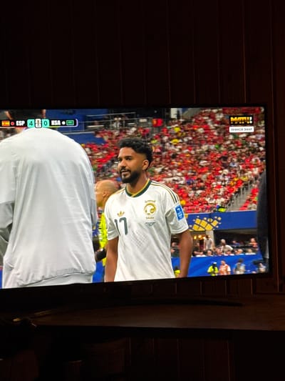 The image shows a television screen displaying a soccer match. The score on the screen indicates that Spain (ESP) is playing against Saudi Arabia (KSA), with the score at 4-0. A player in a white jersey with green trim and the number 17 is visible in the foreground, facing to the right. The jersey has Arabic writing and a Saudi Arabian crest. The background shows a stadium filled with spectators wearing red and white. A "МАТЧ!" logo and "ПРЯМОЙ ЭФИР" (Live broadcast) are visible in the top right corner of the screen. The TV is placed on a dark wooden surface, possibly a piece of furniture. The overall setting suggests a room in Bykovo, Russia, where the match is being watched.