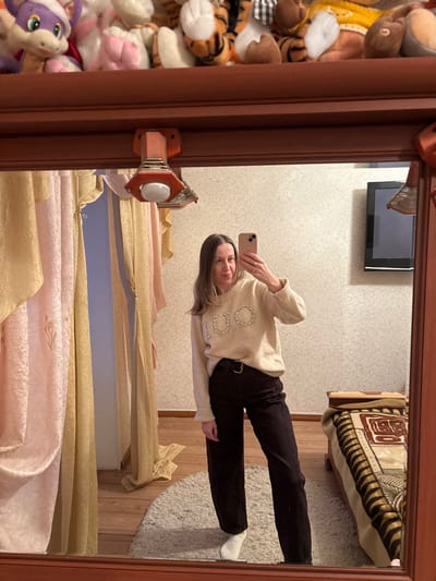 The image displays a mirror selfie, with the entire photograph rotated 90 degrees clockwise, so the subject appears to be standing sideways.
In the center of the mirror's reflection, a woman is visible, appearing to be standing upright in relation to her surroundings. She is captured from her upper chest to her feet. She has long, straight, dark blonde or light brown hair that falls over her shoulders. She is holding a light-colored smartphone (possibly rose gold or beige) in her right hand, angled to capture her reflection. She looks directly at the camera with a gentle smile.
The woman is dressed in a cozy, light beige or cream-colored long-sleeved sweater. The front of the sweater is adorned with shiny embellishments, likely rhinestones or small beads, forming letters. The letters "JUO" are clearly visible, possibly part of a brand name like "LIU JO". She is also wearing dark-colored pants, which appear to be black or very dark brown jeans or trousers, secured with a black belt. Her feet are clad in white socks.
The mirror itself has a thick, dark brown wooden frame. On the viewer's left side of the frame, there's a reddish-orange or reddish-brown bracket-like mechanism with a white circular cap, possibly part of the mirror's mounting or a decorative accent.
The reflected background behind the woman reveals elements of a room. The wall is covered with a light-colored wallpaper featuring a subtle, repeating patterned motif, possibly floral or leafy. In the upper right corner of the reflection, a black-framed television screen is mounted on the wall, appearing to be off. Below and to the right of the TV, a bed is visible, covered with a patterned bedspread or blanket in shades of brown and beige, featuring a geometric design. On the floor, to the right of the woman, there's a round, light-colored (off-white or cream) rug or mat. The rest of the floor is wooden. Below the woman's feet, the reflection shows a pair of drapes or curtains, which are drawn to the side. They consist of a lighter, sheer inner layer in a yellow or gold hue, and a textured outer layer in a soft pink or peach color.
Outside the mirror frame, on the left side of the image, a large collection of stuffed animals or plush toys is piled up. These include several tiger-striped toys, a yellow crocheted item, various white, brown, and gray plush figures, and a distinctive purple creature with large eyes and a green snout at the very bottom left. A black and white checkered fabric item is also visible among the toys.
The scene is lit by what appears to be indoor artificial light. The visible text in the image is "JUO" on the woman's sweater.