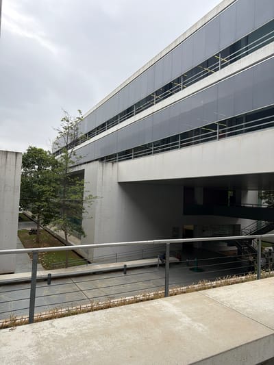 The image shows a modern building in Burjassot, Spain, characterized by its large glass facade and white concrete structure. The building's design incorporates open spaces and landscaping, as evidenced by the trees near its base and the greenery visible in the courtyard. The view is from a low angle, looking up at the building. The weather appears overcast. A metal railing and concrete wall frame the right side of the image, providing a foreground element.