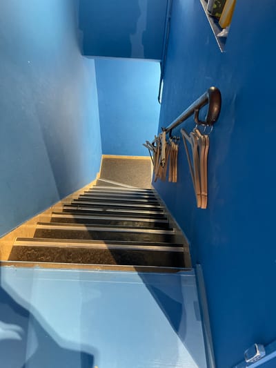 A downward-facing view of a set of stairs in a bright blue, indoor space. The stairs have dark treads with silver nosing. A dark metal handrail runs along the right-hand wall, which is also painted bright blue. Several wooden coat hangers are hanging from the handrail. The scene is clean and well-lit, with shadows cast on the walls and stairs. There are no people or other objects visible. The location is not specified in the image itself, and is thus unknown.