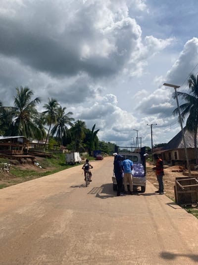 The image shows an outdoor scene in Ufueseni, Nigeria. A paved road runs through the center, flanked by palm trees and low-lying vegetation. A small truck is parked on the side of the road, with several people standing around it. The truck is carrying what appear to be clear plastic containers, possibly filled with water. One person is on a bicycle riding down the road. The sky is overcast with heavy clouds. Buildings and other structures are visible in the background, along with power lines running overhead. The overall impression is of a typical day in a rural or semi-rural area of Nigeria.