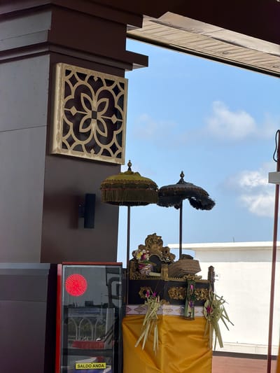 This image depicts an outdoor scene, likely a temple or cultural site in South Denpasar, Indonesia. The sky is visible, suggesting daytime with some clouds. The foreground is dominated by dark brown architectural elements and a decorative lattice panel with a floral motif.
In the mid-ground, traditional Balinese offerings are prominently displayed. These include ornate umbrellas, one dark and fringed, and another in a muted green with decorative tassels, both appearing to be part of a procession or ceremony. They are placed on a raised platform draped with a vibrant yellow cloth, adorned with gold trim and what appear to be woven palm leaf decorations. The overall arrangement suggests a religious or ceremonial context.
Further to the right, a glass-fronted display case is visible. Inside, a red circular pattern is illuminated, possibly an indicator or part of an exhibit. A yellow sticker with text is affixed to the right side of the display case. The text on the sticker is "SALDO ANDA", which translates from Indonesian to "YOUR BALANCE". This hints at the possibility of a transactional or service element within the cultural setting, perhaps related to donations or ticket sales.
The architecture suggests a blend of modern and traditional elements, with the dark brown structures contrasting with the bright yellow fabric and the intricate lattice work. The lighting indicates it is likely daytime, with diffused sunlight illuminating the scene. The presence of the ornate umbrellas and offerings strongly points to a cultural event or ongoing religious practice.