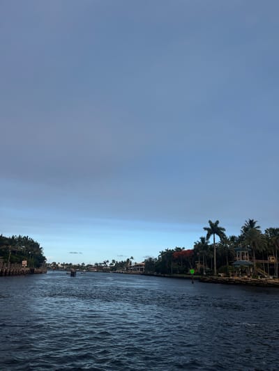 The image depicts a wide, dark blue canal in Fort Lauderdale, Florida, with the viewer positioned on the water, looking towards the horizon. The canal is lined on both sides by lush green vegetation, primarily palm trees, and luxurious waterfront homes with ornate architecture. Several boats are visible on the water, some in the distance, suggesting recreational activity. The sky above is a pale blue, transitioning into a hazy gray towards the horizon, indicating either early morning or late afternoon light. The overall scene conveys a sense of tranquility and opulence, characteristic of Fort Lauderdale's affluent canal communities.