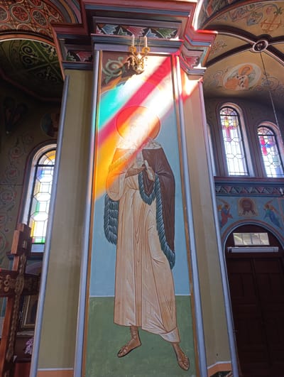 The photograph captures the interior of a church in the city of Kitskany, Moldova. The central focus is a large, rectangular pillar adorned with a fresco depicting a saint. The saint is rendered in traditional religious iconography, wearing a pale yellow robe and a dark brown cloak trimmed with fur. A halo encircles the figure's head. Diagonal rays of light, colored red, orange, yellow, and green, stream across the fresco from an unseen source, likely stained-glass windows, illuminating the saint and casting colorful shadows.

The pillar is framed by an arched ceiling painted with intricate patterns and religious imagery. To the left, a stained-glass window with geometric patterns allows sunlight to enter the building. A wooden cross, ornately carved, stands to the left of the pillar. In the background, on the right side of the image, a large wooden door is visible, set within an arched doorway. Above the door, another section of the wall is painted with religious figures, including angels. The overall environment suggests a place of worship, characterized by religious art and the play of light.