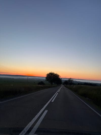 The image shows a road with a white double line down the center, stretching into the distance. The road is lined with dry grass and scattered trees. In the background, a hazy landscape is visible under a gradient sky that transitions from a deep blue at the top to a bright orange and red near the horizon, indicating either sunrise or sunset. The location is identified as the city of Pașcani, Romania. There are no people or any discernible action occurring in the image. The environment appears rural and tranquil.