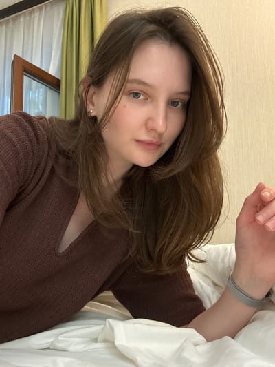 This media file features a young woman lying in bed. She has long, brown hair that is casually styled and falls around her face and shoulders. She is wearing a brown, textured sweater. Her eyes are open, and she appears to be looking towards the camera. A silver mesh watch is visible on her left wrist.
The setting appears to be indoors, likely a bedroom, with white bedding prominently featured. A window with green curtains and a view of what looks like a window frame with a reflection is visible in the background. The lighting suggests it could be daytime, possibly morning or afternoon. There is no visible text in the image. The overall mood is relaxed and intimate, as if capturing a candid moment.