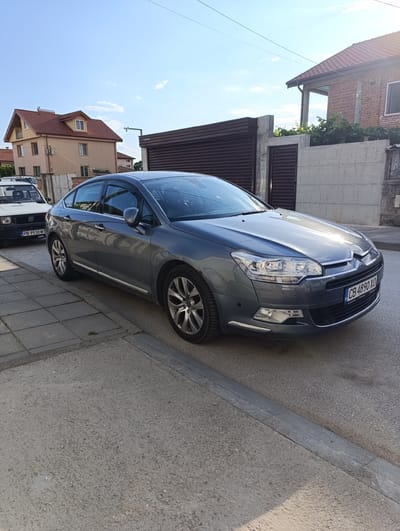 The image captures a sunny outdoor scene on a residential street in Rakovski, Bulgaria, featuring two parked cars and several houses.
In the foreground and centrally located is a dark gray (or perhaps a deep blue-gray) Citroën C5 sedan (second generation), parked on the asphalt road next to a paved sidewalk. The car is angled slightly towards the right, with its front bumper and headlight area prominently visible. The sun reflects brightly off its hood and windshield. Its Bulgarian license plate reads "CB 4890 XO," indicating registration in Sofia. Small text "www.bomba.bg" is partially visible on the license plate holder.
To the left and partially obscured by the Citroën is a white first-generation Fiat Panda, also parked on the street. Only its front and side are visible, including its Bulgarian license plate, which reads "PB 9930 MT," indicating registration in Plovdiv.
The background features several residential buildings under a clear, bright blue sky with a few faint clouds. On the left, a multi-story house with light beige walls and a prominent reddish-brown tiled gabled roof, featuring an attic dormer window, is visible. To the right, another building with a similar tiled roof is partially seen, appearing to be constructed of brick. A tall concrete wall with dark brown slatted garage doors or gates separates the street from some of the properties. Green foliage can be seen peeking over these walls.
Overhead, thin power lines or communication cables crisscross the sky. A security camera on a pole extends from one of the buildings towards the center. The road surface is asphalt, and the sidewalk consists of light grey concrete pavers.
The scene is illuminated by bright daylight, suggesting late morning or afternoon, with strong shadows cast by the vehicles and buildings. No people or specific activities are visible, conveying a calm, everyday street ambiance.