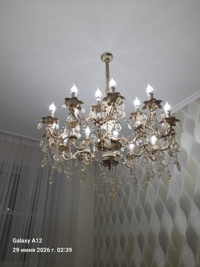 A chandelier with numerous crystal accents hangs from a white ceiling. The chandelier features a metallic gold structure with multiple arms, each holding a candle-shaped light bulb. White sheer curtains are visible on the left side of the image, and a wall with a grey and white wave pattern is on the right. The bottom left corner of the image contains text that reads "Galaxy A12" and "29 июня 2026 г. 02:39". The image is taken in Kara-Köl City, Kyrgyzstan.