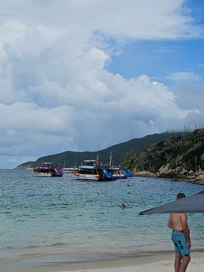 The image displays a coastal scene at a beach in Arraial do Cabo, Brazil. In the immediate foreground, on the right, a shirtless man with grey hair stands on light beige sand, wearing blue swim trunks with a red floral and black sea turtle pattern. He is partially obscured by a dark grey canopy extending from the right frame. The clear, shallow water beyond the sand is turquoise, showing gentle ripples, with several individuals swimming at various distances from the shore.
In the midground, three multi-level tour boats are anchored. The two most prominent vessels have white hulls; one on the left features a purple upper deck and an attached purple slide descending into the water, while the boat on the right displays a blue upper deck with a corresponding blue slide. Both boats are populated with numerous passengers. A smaller, white boat is partially visible between these two.
The background on the right is dominated by a green hill or mountain, its slopes covered in dense vegetation, including numerous tall, slender cacti and several trees appearing bare of leaves. Rocky outcrops are visible at the base of the hill where it meets the water. The sky above is largely filled with large white and grey cumulus clouds, with patches of clear blue sky visible, particularly in the upper right quadrant, where a few small, dark bird-like shapes are in flight.