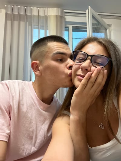 An indoor, eye-level selfie captures two individuals, a man and a woman, in an affectionate pose within a residence in Karavelovo, Bulgaria. The man, positioned on the left, has short dark hair with faded sides, and is wearing a light pink crew-neck t-shirt. He is seen leaning in to kiss the woman's right cheek. The woman, on the right, has light brown hair and is wearing black-framed eyeglasses. Her eyes are closed, and her right hand is gently touching her chin/cheek. She is wearing a white tank top, revealing a silver necklace with a heart-shaped pendant inscribed "love" and a small round charm. In the background, white pleated curtains are visible on the left, and a white-framed window is partially open on the right, showing a clear blue sky outside.