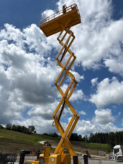 The image depicts a tall, yellow scissor lift in the foreground, extending upwards against a blue sky with fluffy white clouds. Two construction workers, wearing orange safety vests, are visible on the elevated platform of the scissor lift. The scissor lift itself is a complex mechanical structure with interconnected metal beams forming a crisscross pattern. In the background, a green, hilly landscape is visible, with trees dotting the slopes and a winding road leading upwards. Further in the distance, a dense forest stands at the top of the hill. A white truck is parked to the right of the scissor lift. The overall scene suggests a construction site in a rural or semi-rural area. While the prompt mentions Janowice, Poland, there is no explicit visual evidence within the image to confirm this specific location. The environment appears to be an outdoor setting, possibly undergoing development or construction, given the presence of heavy machinery and the unfinished appearance of some areas in the mid-ground.